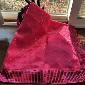 May include: A bright pink scarf with a sequined border. The scarf is made of a sheer fabric and is decorated with small, scattered sequins. The border is a darker shade of pink and is densely covered in sequins. The scarf is draped over a black mannequin hand.