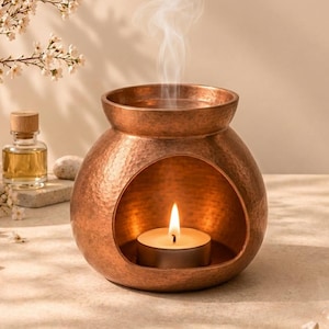 May include: A round, hammered copper oil burner with a wide opening at the front. A small, lit tea light candle sits inside the base, and a thin wisp of white smoke rises from the top bowl. The metallic surface has a textured, dimpled finish.