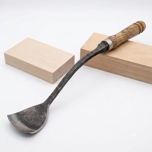 May include: A hand-forged metal tool with a curved, wide, flat-edged blade and a textured dark gray finish. The tool features a long, slender neck and a cylindrical wooden handle with a light brown grain, secured by a silver-colored metal ferrule.