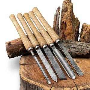 Könnte beinhalten: Ein Set aus fünf Drechselmeißeln mit langen, hellen Holzgriffen und dunklen, geschmiedeten Metallklingen. Jedes Werkzeug hat eine eigene Klingenform, darunter Röhren und ein Flachmeißel, mit einem kleinen dreieckigen Logo auf dem Metall.