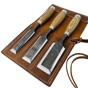 May include: A set of three wood chisels with dark, textured metal blades and light-colored wood handles, arranged in a brown leather tool roll with white stitching and a leather tie closure. The metal blades show a dark, mottled finish.