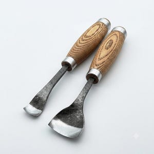 May include: Two hand tools with wooden handles and silver metal ferrules. The tools feature dark, textured, forged metal blades. One blade is narrow and curved, while the other is wider and scoop-shaped, both showing a raw, hammered finish.