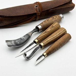 May include: A set of four wood carving tools with textured wooden handles and metal ferrules, featuring various blade shapes including a large curved gouge, a straight detail knife, a hook knife, and a small detail blade, with a leather roll.