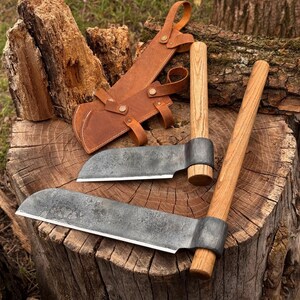 Puede incluir: Dos hachuelas de hierro forjado a mano con mangos de madera clara y una funda de cuero color canela. Las hachuelas cuentan con hojas de metal oscuro curvadas con un acabado texturizado, fijadas firmemente a mangos cilíndricos de madera.