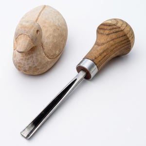 May include: A light-colored wooden duck carving with visible chisel marks and a metal wood carving gouge featuring a rounded wooden handle and a silver-colored steel blade, positioned together on a plain white surface.