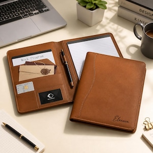 May include: A tan leather portfolio folder featuring a notepad, a black pen, and interior slots for cards and documents. The front cover is embossed with the name Eleanor. The interior includes a paper labelled Work Program and a small card.