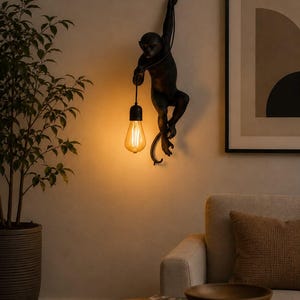 May include: A black monkey-shaped wall lamp featuring a matte finish. The monkey figure is suspended from a black cord and holds a single glowing Edison-style light bulb in one hand, casting a warm, amber-toned light against the wall surface.