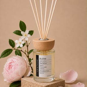 May include: A clear glass reed diffuser bottle with a light wood cap and six wooden reeds. The label reads VENTORE BLOOM 100 ml / 3.4 fl.oz Reed diffuser, peony, blush suede, jasmine, Handcrafted in Oxford, Batch 001. A pink peony and white flowers.