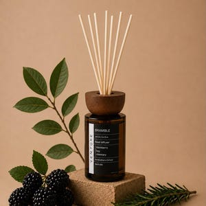 May include: A brown glass reed diffuser bottle with a dark wooden cap and six light-coloured reeds. The black label reads BRAMBLE, 100 ml / 3.4 fl.oz, Reed diffuser, blackberry, bay, rosemary, Handcrafted in Oxford, Batch 001. Includes blackberries and rosemary.