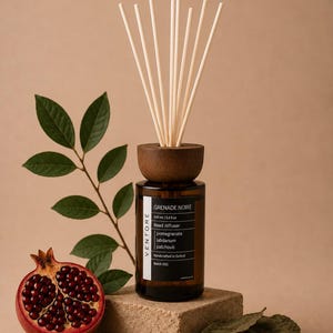May include: A brown glass reed diffuser bottle with a dark wooden cap and white reeds. The label reads GRENADE NOIRE, 100 ml / 3.4 fl. oz, Reed diffuser, pomegranate, labdanum, patchouli, Handcrafted in Oxford, Batch 001. A pomegranate and leaves are nearby.