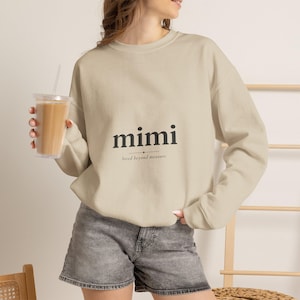 以下が含まれることがあります： 胸の中央に黒い文字でmimi、その下に小さくloved beyond measureとプリントされたベージュのクルーネックトレーナー。長袖でリブ編みの襟元が特徴で、グレーのデニムショートパンツと組み合わされています。