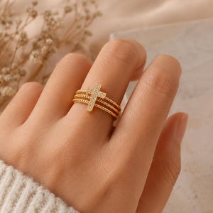 Könnte beinhalten: Ein goldfarbener Ring bestehend aus drei übereinanderliegenden, gedrehten Bändern, die durch ein zentrales vertikales Kreuz verbunden sind. Das Kreuz ist mit kleinen, klaren, funkelnden Steinen besetzt, was für eine strukturierte Optik sorgt.