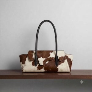 Könnte beinhalten: Eine strukturierte Handtasche mit braun-weißem Kuhfellmuster und strukturierter Felloberfläche. Die Tasche hat einen horizontalen, rechteckigen Körper mit Klappverschluss und zwei hohen, dünnen Tragegriffen aus schwarzem Leder.