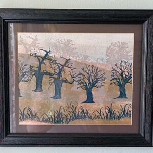 May include: A framed print featuring a landscape with dark blue silhouettes of bare trees and tall grasses against a background of muted tan and light brown hills. The artwork is enclosed in a textured black wooden frame with a dark brown mat.