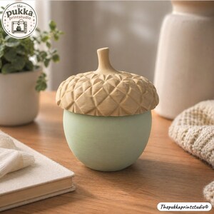 May include: A decorative acorn-shaped container featuring a smooth, light sage green base and a textured, tan-coloured cap with a geometric, diamond-patterned surface. The lid is topped with a small, rounded stem, creating a rounded silhouette.