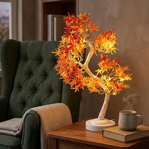 Puede incluir: Una lámpara decorativa de árbol LED con hojas de estilo arce en tonos naranja y rojo vibrantes, iluminadas por luces cálidas. El árbol presenta un tronco blanco texturizado y se asienta sobre una base circular blanca para una mesa auxiliar.