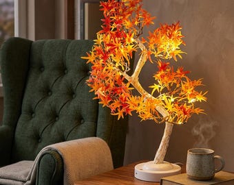 May include: A decorative LED tree lamp featuring vibrant orange and red maple-style leaves illuminated by warm internal lights. The tree has a textured white trunk and sits on a circular white base, providing a glowing accent for a side table.