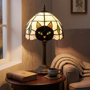 May include: A stained glass table lamp featuring a cream-colored dome shade with a black cat face design, green eyes, and an orange nose. The lamp has a dark bronze-toned metal base and emits a warm glow through the glass panels.