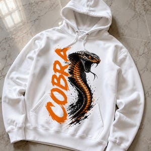 Könnte beinhalten: Ein weißer Kapuzenpullover mit einem Grafikdruck einer Kobra mit orangefarbenen und schwarzen Schuppen. Das Wort COBRA ist in großen, fetten, orangefarbenen Pinselstrich-Buchstaben links neben der Schlange mit offenem Maul aufgedruckt.