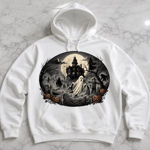 May include: A white hooded sweatshirt featuring a circular graphic on the chest. The illustration depicts a haunted mansion, a ghost, a witch, a scarecrow, and jack-o'-lanterns in a graveyard setting under a full moon with dark, moody tones.