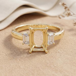 May include: A gold ring setting featuring a central rectangular prong mount designed for a stone, flanked by two clear emerald-cut side stones. The metal band is polished yellow gold, and the text DOMIXA is visible above the central setting.