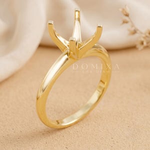 May include: A polished yellow gold ring setting featuring a four-prong head designed to hold a single gemstone. The band is smooth and rounded, and the metal surface reflects light. The text DOMIXA LAB DIAM is visible on the ring shank.