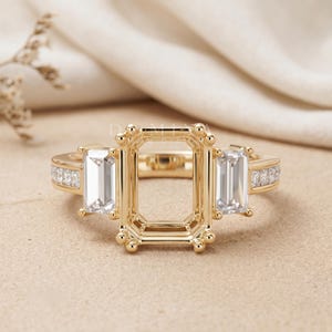 May include: A gold ring setting featuring a large rectangular center prong setting flanked by two baguette-cut clear stones. The band is accented with small pavé-set stones along the sides. The text DEMIYA is visible above the center setting.