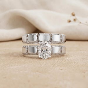 May include: A silver-toned ring set consisting of two bands. The top band features a repeating rectangular pattern. The bottom band features a central oval-cut clear stone set against a matching rectangular patterned band. The metal has a polished finish.