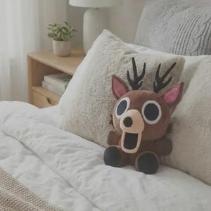 May include: A brown plush deer toy with large circular eyes, a light tan muzzle, a wide open mouth, and black antlers. The stuffed animal features pink inner ears and dark brown paws, sitting upright with a soft, rounded body and a cartoonish appearance.