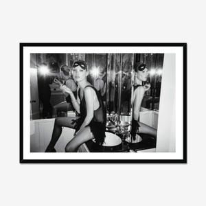 Può includere: Una fotografia in bianco e nero in una cornice nera che ritrae una persona che indossa una maschera scura e un abito senza maniche con frange. La persona è seduta davanti a uno specchio, con riflessi visibili nel vetro e bagliori di luce.
