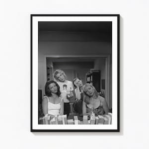 May include: A black and white framed photograph featuring four individuals positioned behind a row of books. The subjects are wearing casual tops, including tank tops and a graphic t-shirt, and are arranged in a group setting within an interior space.