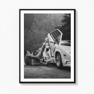 May include: A black and white photographic print featuring a person lying on the ground next to a white sports car with an open scissor door. The person is wearing casual clothing and has one leg extended upward, resting against the car door.
