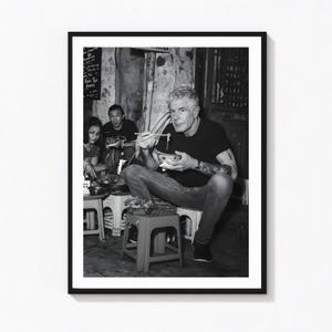 Könnte beinhalten: Ein Schwarz-Weiß-Foto zeigt eine Person mit kurzem Haar in einem dunklen T-Shirt und Jeans, die auf einem kleinen Kunststoffhocker sitzt und mit Stäbchen aus einer Schüssel isst. Eine weitere Person sitzt in der Nähe an einem Tisch.