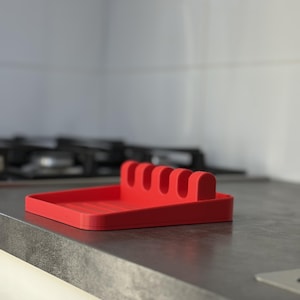 May include: A bright red silicone kitchen utensil rest featuring a flat rectangular base with raised ridges and a back support section containing four distinct slots designed to hold cooking tools in an upright or horizontal position.
