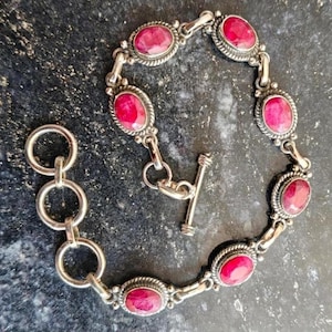 May include: A silver chain bracelet featuring six oval-shaped, deep pink cabochon stones set in twisted metal bezels. The bracelet includes a toggle clasp mechanism and a series of three circular silver links for adjustable sizing.
