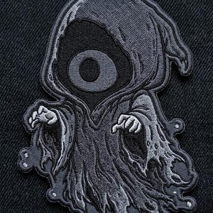 May include: An embroidered patch featuring a stylized, dark gray hooded figure with a single large, circular eye in the center of its face. The figure wears a tattered, flowing robe with visible stitching details and has two hands reaching forward.