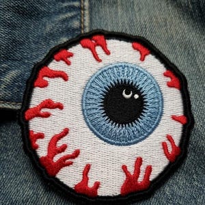 May include: A circular embroidered patch featuring a realistic human eyeball design. The patch has a white sclera with red bloodshot veins, a light blue iris, and a black pupil with a small white highlight, all bordered by a thick black edge.