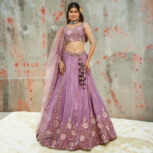 May include: A lavender lehenga choli set featuring a sleeveless blouse and a floor-length skirt, both adorned with intricate floral embroidery and beadwork. The ensemble includes a matching sheer dupatta with a decorative border and tassel details.
