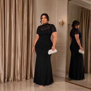 May include: A floor-length black gown featuring a high neckline, short sleeves, and a sheer, vertically striped yoke panel. The dress has a fitted bodice that flares slightly toward the hem and is paired with a small, textured white clutch purse.
