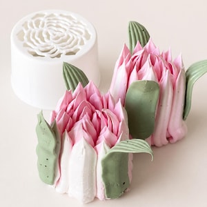 May include: Two piped frosting flowers in shades of pink and white with green leaves, positioned next to a white plastic circular piping nozzle featuring a detailed floral cutout pattern on its top surface for creating intricate icing designs.