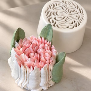 Könnte beinhalten: Ein dekoratives Objekt in Blumenform mit weißen und rosafarbenen, wie Zuckerguss geformten Blütenblättern und salbeigrünen Blättern, begleitet von einem zylindrischen weißen Behälter mit einem komplexen, lasergeschnittenen Blumenmuster.