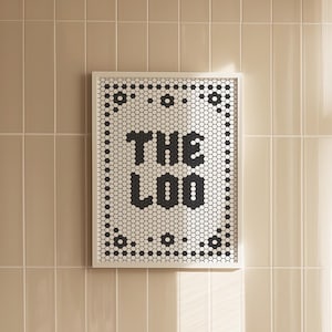May include: A framed black and white mosaic art piece with the text "THE LOO" hangs on a beige tiled wall above a white bathtub. Sheer white curtains are to the right, and a beige towel rests on the tub.