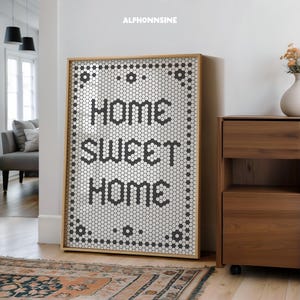 Puede incluir: Un cuadro enmarcado con las palabras "HOME SWEET HOME" en negro, con forma de hexágono. El fondo es blanco con un patrón de pequeñas baldosas hexagonales en blanco y negro. El marco es de madera clara.