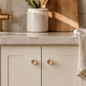 May include: Two circular, light-colored wooden cabinet knobs with a concentric ring texture mounted on off-white kitchen cabinet doors. The knobs feature a small, recessed center point and a smooth, rounded profile, providing a functional grip.
