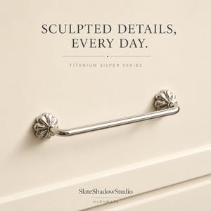 Può includere: Una maniglia per cassetto in metallo color argento con una barra cilindrica liscia e basi di montaggio decorative a forma di fiore. Il testo in alto recita SCULPTED DETAILS, EVERY DAY. TITANIUM SILVER SERIES, in basso SlateShadowStudio HARDWARE.
