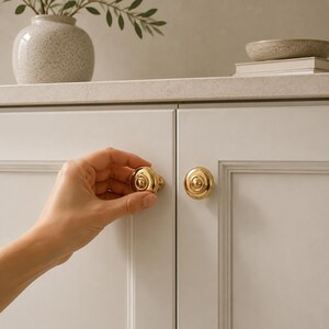 May include: A round, polished gold cabinet knob with a tiered, concentric circular design and a beaded outer edge. The knob is mounted on a light gray cabinet door with a recessed panel frame, featuring a smooth, reflective metallic finish.