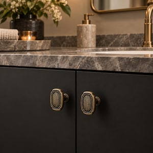 May include: Two rectangular cabinet knobs with a brass finish and a textured, cross-hatched center pattern. The knobs are mounted on matte black cabinet doors, featuring a raised border and a central grid design that provides a tactile surface.