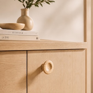 May include: A light-colored wooden cabinet door featuring a circular, recessed wooden knob. Above the cabinet, two stacked books are visible, with the top book titled KINFOLK and the bottom book titled at 100, alongside a small wooden bowl and vase.