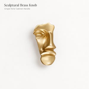 Puede incluir: Un pomo de latón de color dorado con un diseño de rostro escultórico abstracto. El texto "Sculptural Brass Knob" y "Single Hole Cabinet Handle" está impreso sobre el objeto. El pomo tiene un acabado metálico liso con contornos curvos y orgánicos.