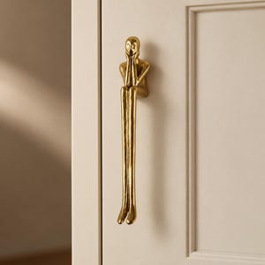 May include: A gold-toned metal cabinet handle shaped like a stylised human figure sitting with legs extended downwards and hands resting near the face. The figure has a smooth, reflective surface and is mounted vertically on a light-coloured door panel.